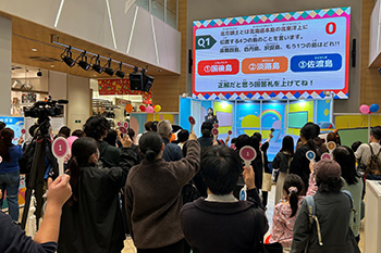 親しみやすい雰囲気のセットが組まれ、司会者がクイズを出題している、クイズの内容を観衆が眺めている