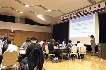 スクリーンに映し出された資料を、机に座った人々が真剣に見ている様子